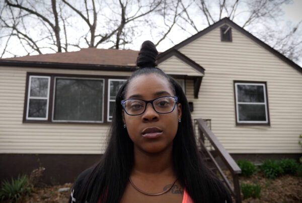A woman with glasses and a top bun standing outside in front of a house beneath trees and a gray sky