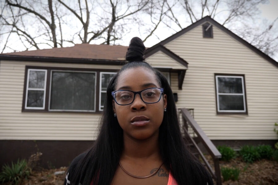 A woman with glasses and a top bun standing outside in front of a house beneath trees and a gray sky