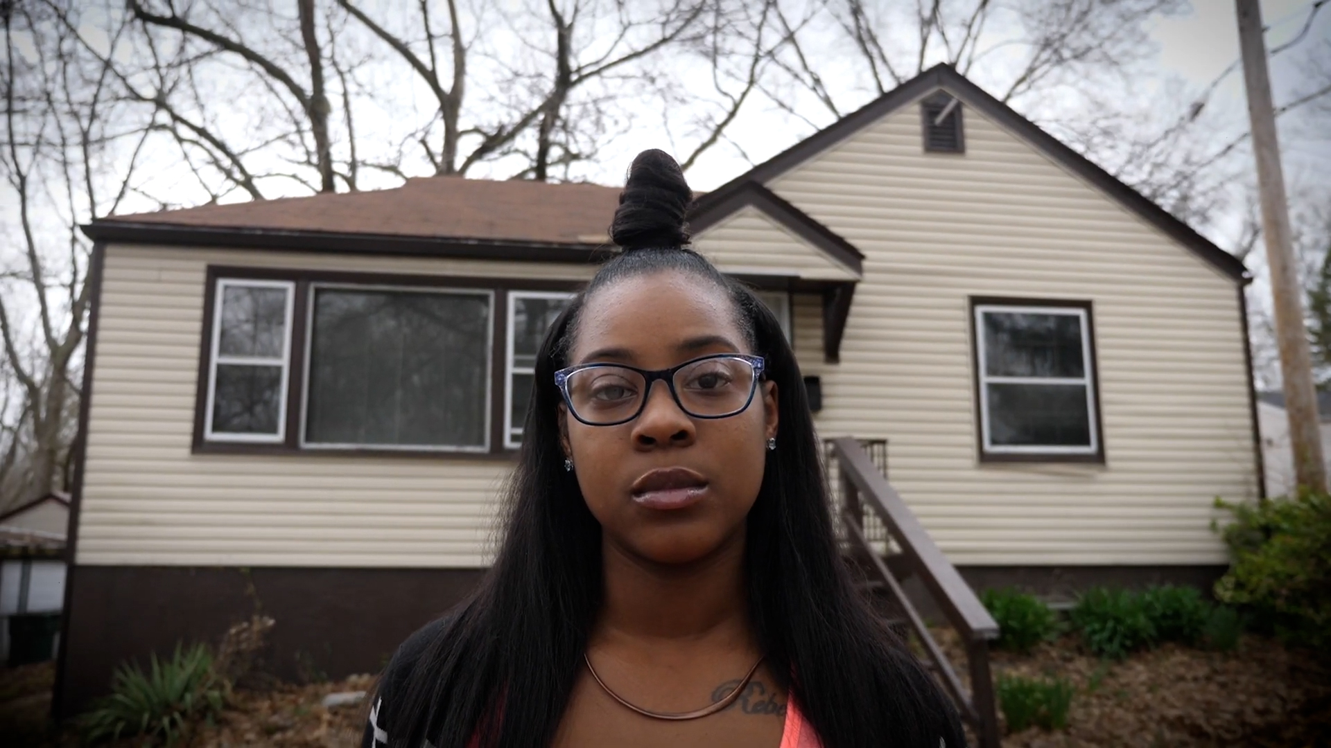 A woman with glasses and a top bun standing outside in front of a house beneath trees and a gray sky