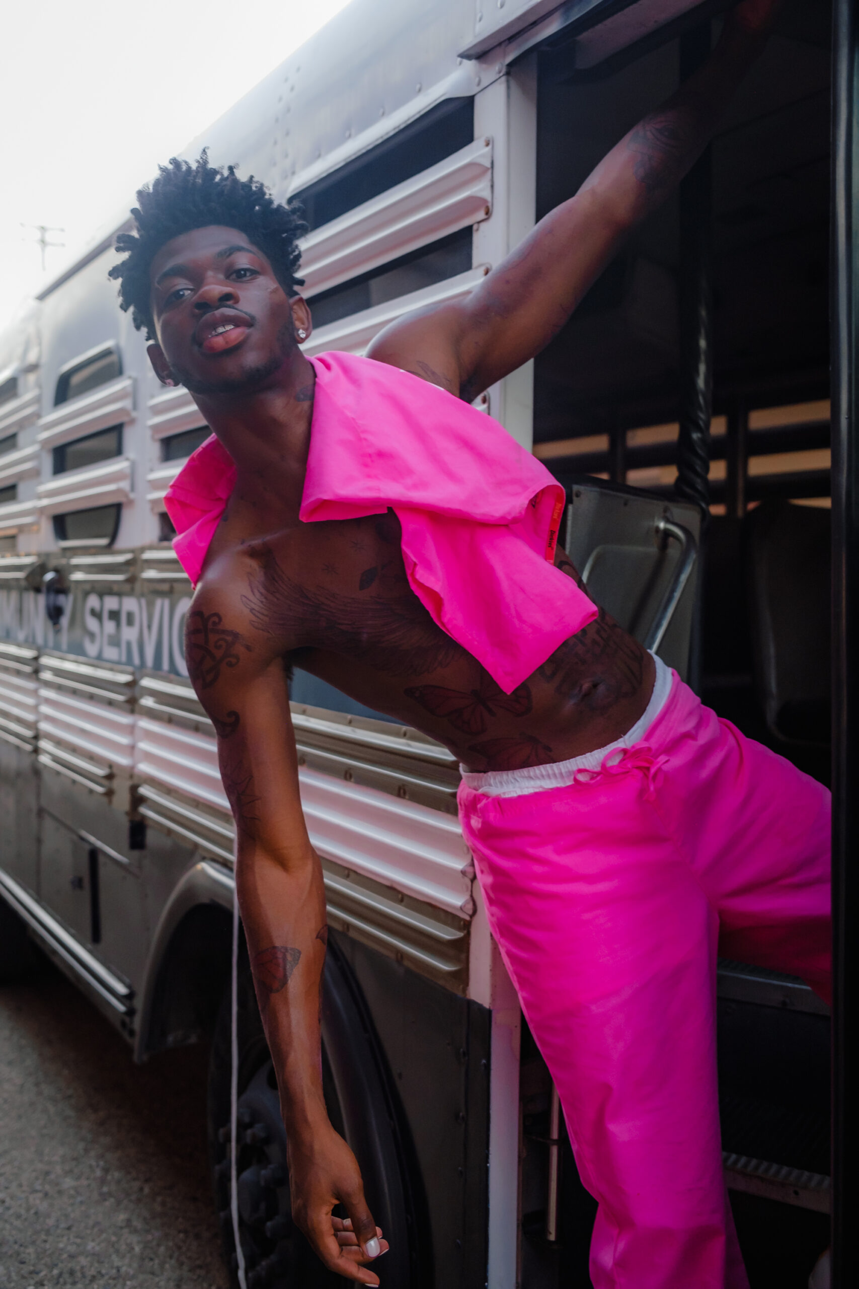 Lil Nas X, shirtless with a bright pink shirt over his shoulder and wearing bright pink shorts, leaning on the side of a train looking into the camera; credit Adrian Per