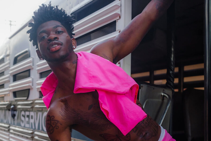 Lil Nas X, shirtless with a bright pink shirt over his shoulder and wearing bright pink shorts, leaning on the side of a train looking into the camera; credit Adrian Per