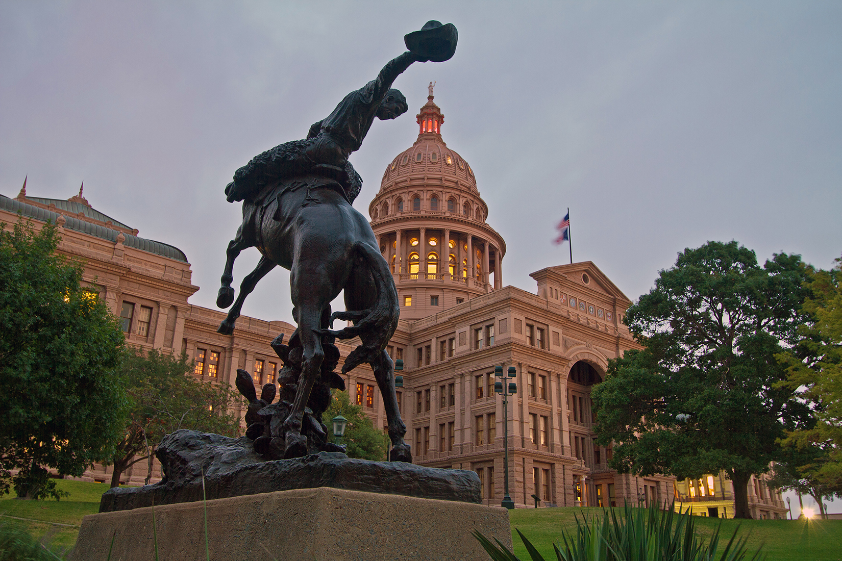 A statue of a man riding a horse with a cowboy hat in his hand in front of a capitol building