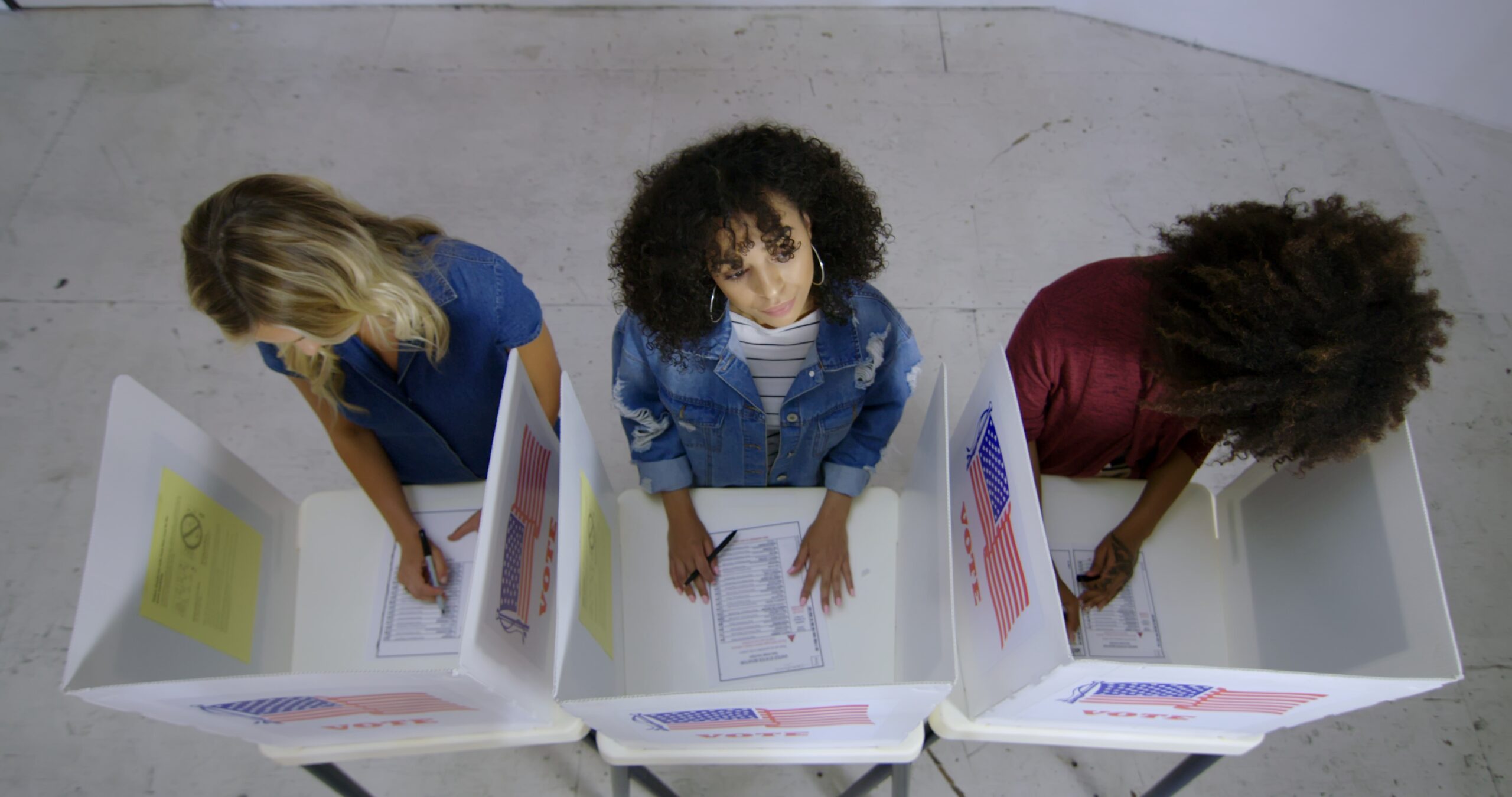 a birds-eye-view of a group of three people at ballot boxes, one in thought