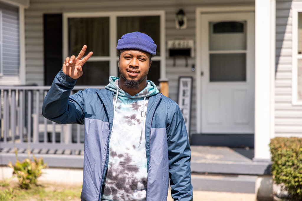 St. Louis Client standing in the sun, with a hat and jacket, looking into the camera holding up a "peace sign" with his fingers