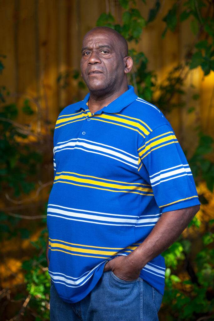 Houston client, Hervis, standing outside in the shade amongst greenery, in a striped polo and blue jeans