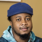 a man with a beard and blue beanie looking into the camera in front of a green wall