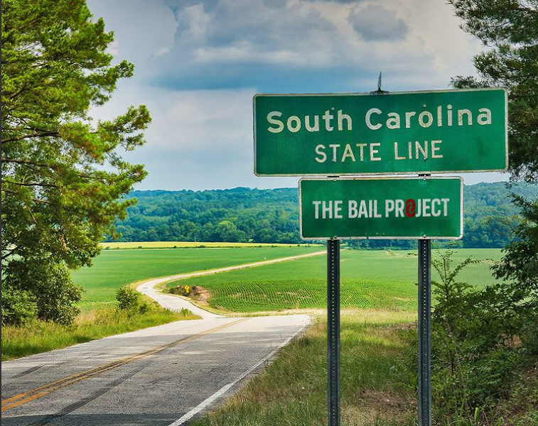 a green street sign sitting on the side of a road, reading "South Carolina, State Line", followed by The Bail Project logo
