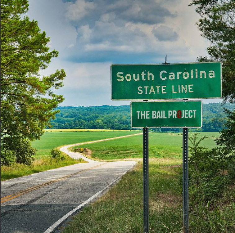 a green street sign sitting on the side of a road, reading "South Carolina, State Line", followed by The Bail Project logo