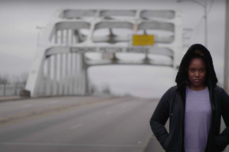 A woman with her hood up, shadowed, standing outside by a bridge, staring determinedly into the camera, discusses the cash bail system