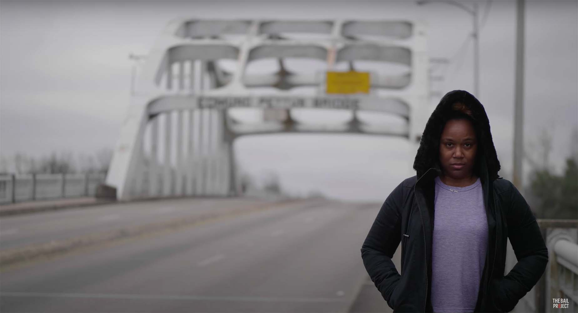 A woman with her hood up, shadowed, standing outside by a bridge, staring determinedly into the camera