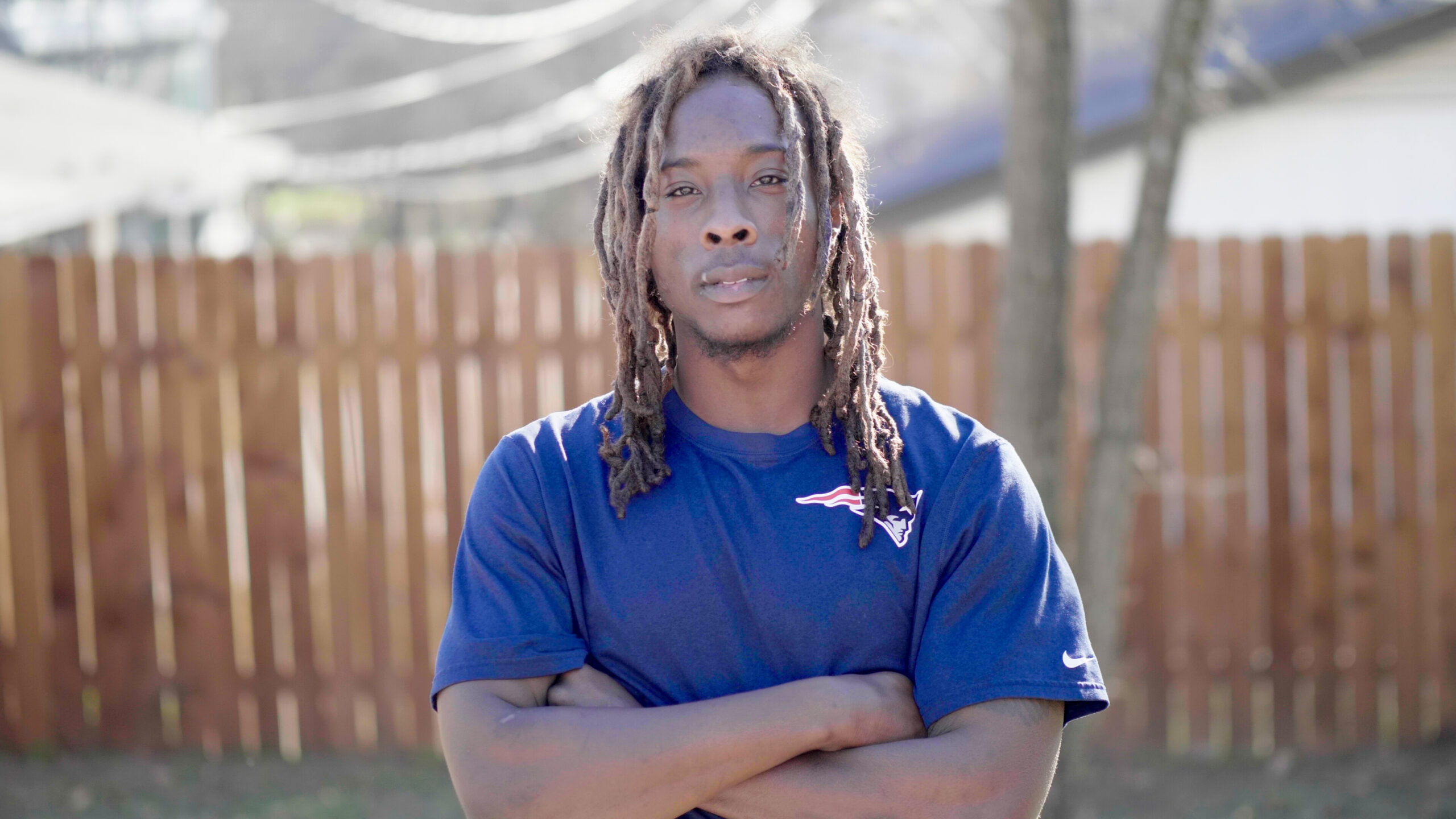 A man with dreadlocks standing in front of a fence with arms across his chest