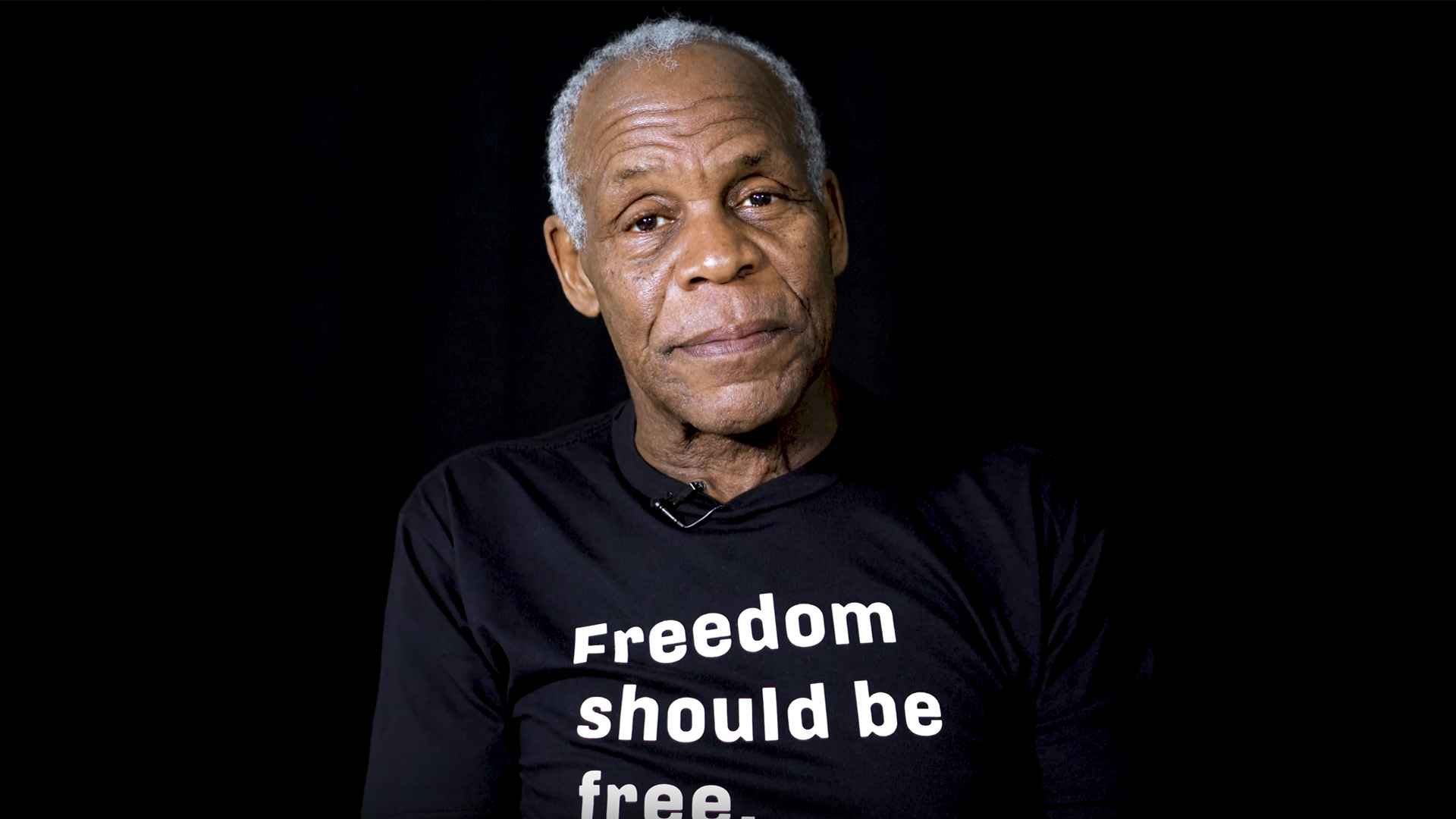 Danny Glover with a "Freedom should be free" shirt staring into the camera, against a black background