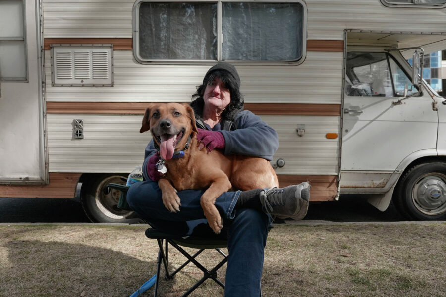 A person in a beanie with long black hair, sitting with a dog in front of a mobile home.