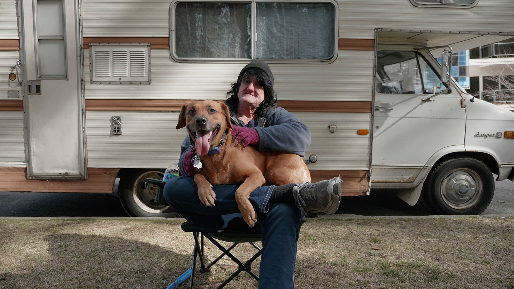 A person in a beanie with long black hair, sitting with a dog in front of a mobile home.