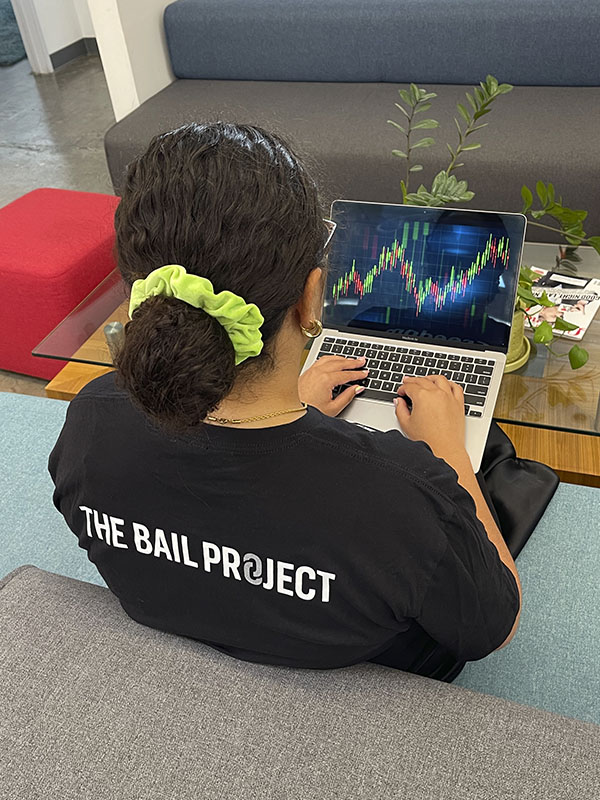 a woman on a couch in a "Bail Project" shirt, using a laptop computer.