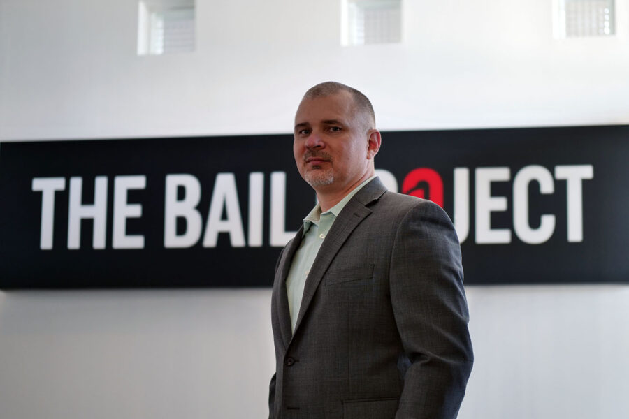 a man, Bail Project CEO David Gaspar, in a suit standing in front of a Bail Project Logo sign.