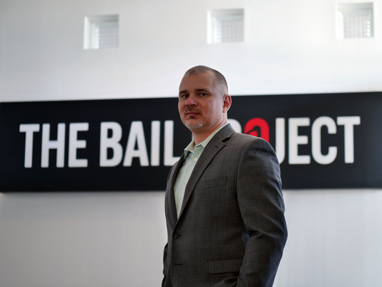 a man, Bail Project CEO David Gaspar, in a suit standing in front of a Bail Project Logo sign.