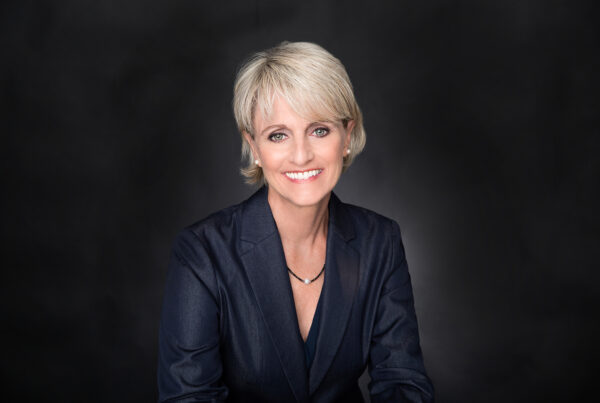 A white woman against a black background, with blond hair in a blazer smiling and looking into the camera